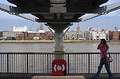 Girl in jeans walking under the Millennium Bridge in London, England, with the opposite side of the River Thames in sunshine in the backgound